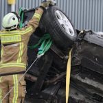 Firefighter in protective gear secures overturned car at an accident scene with ropes and equipment.