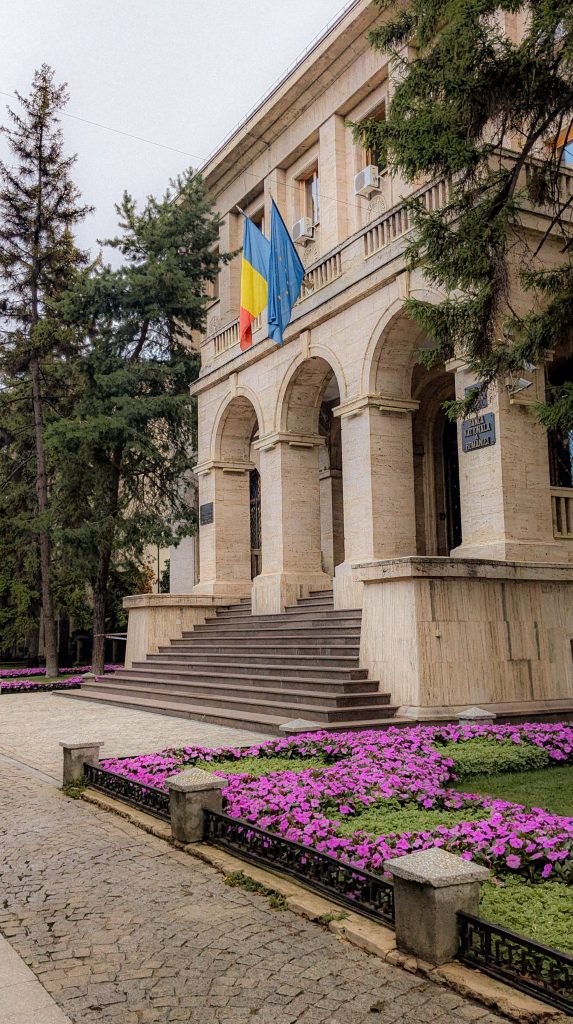 Historic building with flags and colorful flower beds in a lush garden setting.